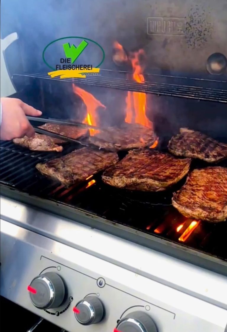 Fleischstücke brutzeln auf einem Grill, während Flammen und Rauch aufsteigen.