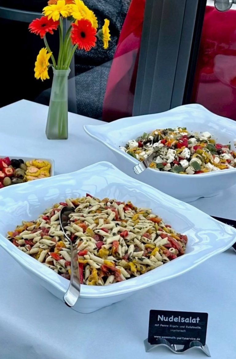 Bunte Nudel- und Gemüsesalate in weißen Schalen, mit Blumenarrangement im Hintergrund.