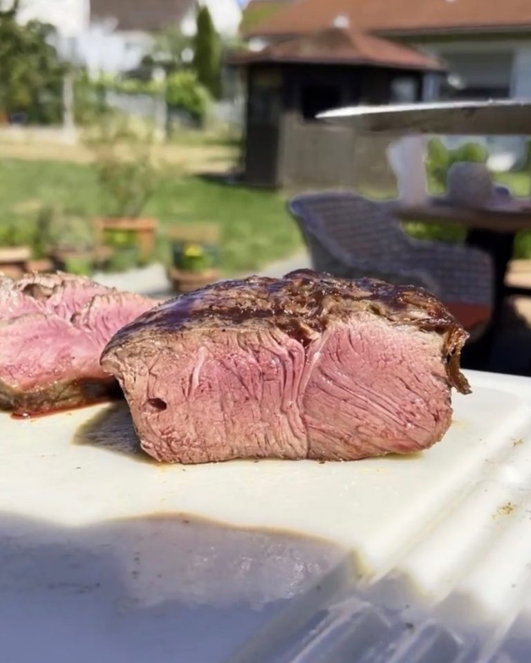 Saftiges, rosa gebratenes Fleisch auf einem Schneidebrett, im Hintergrund Gartenansicht.